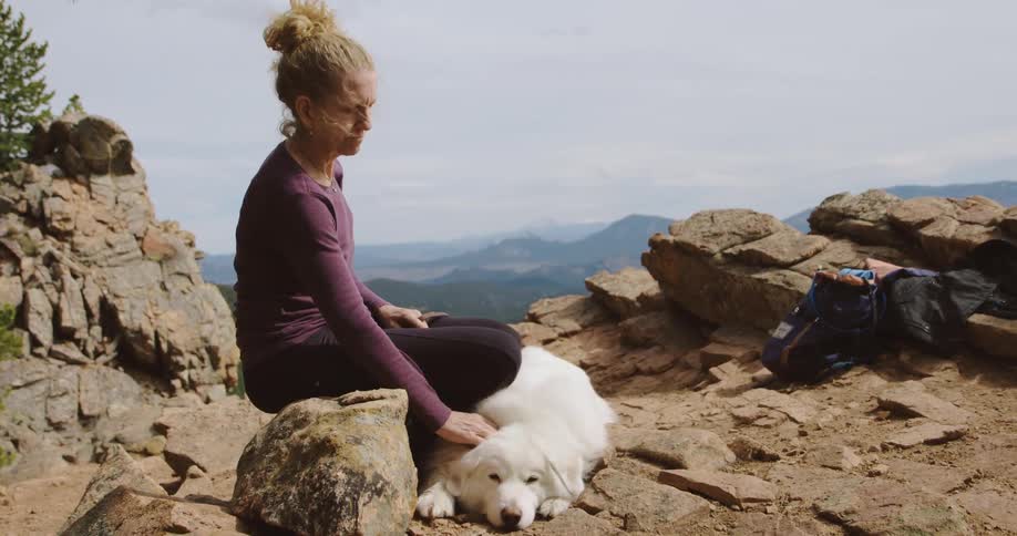 A woman petting a white dog while sitting on rocks in mountains