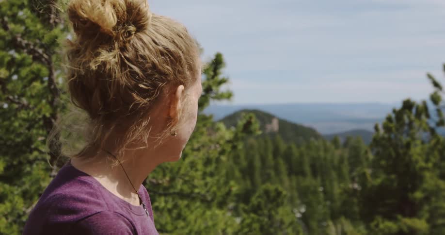 A breeze blows tendrils of a womans hair as she looks out at tree tops and hills