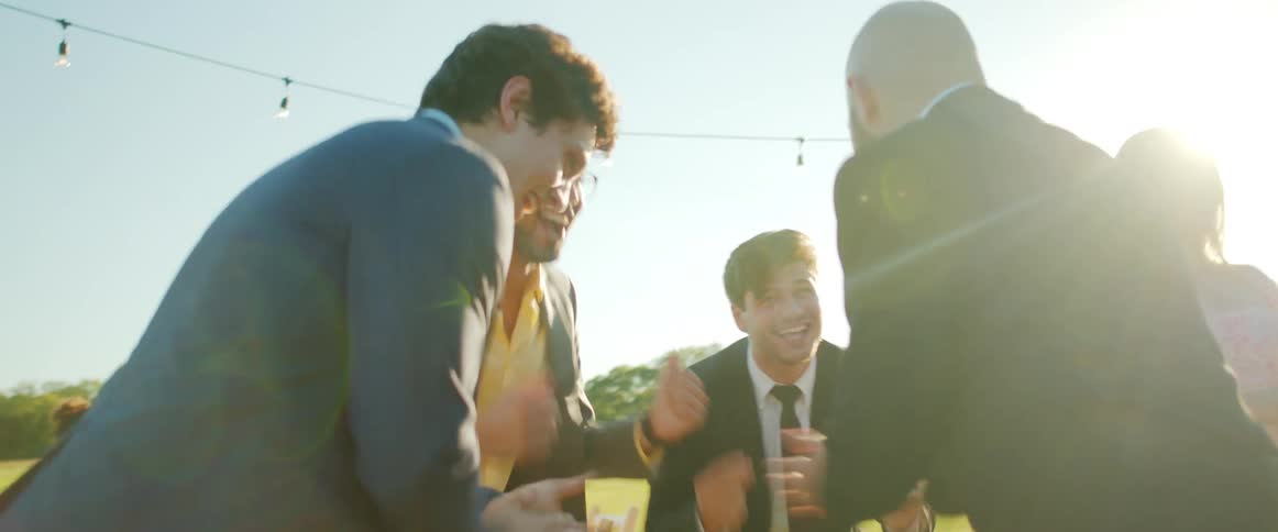 Four men in a circle laugh and clap outdoors on a sunny day