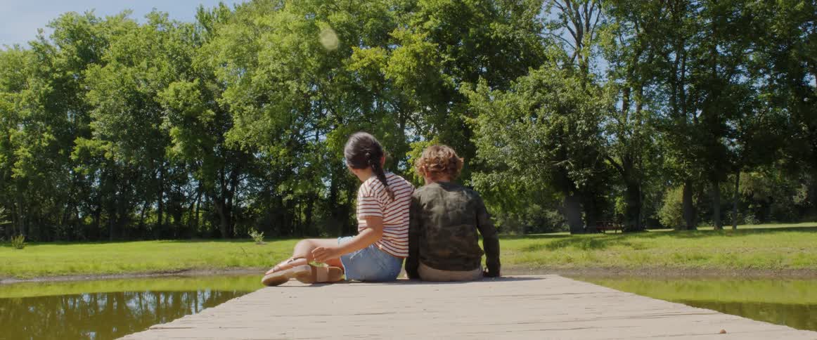 Boy and girl sit on a dock at a pond with trees on the other side
