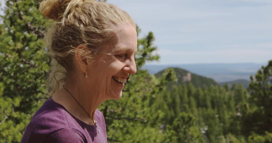 Happy blonde woman standing in front of a forest in the mountains on a clear sunny day