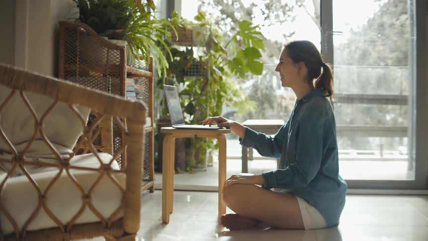 A woman sits on the floor with her laptop and waves at the screen