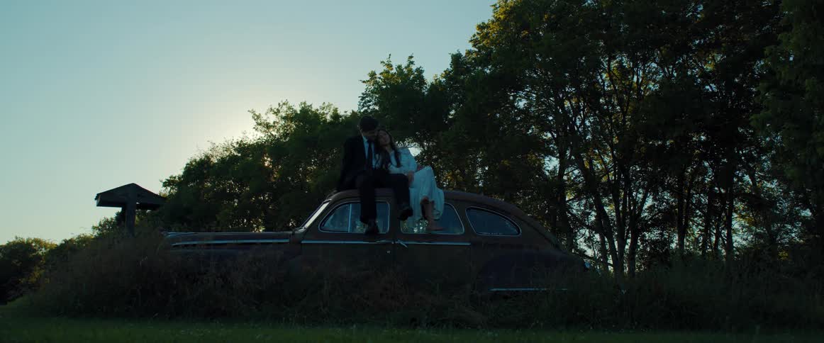 A man hugs his bride as they sit atop a vintage car in a park