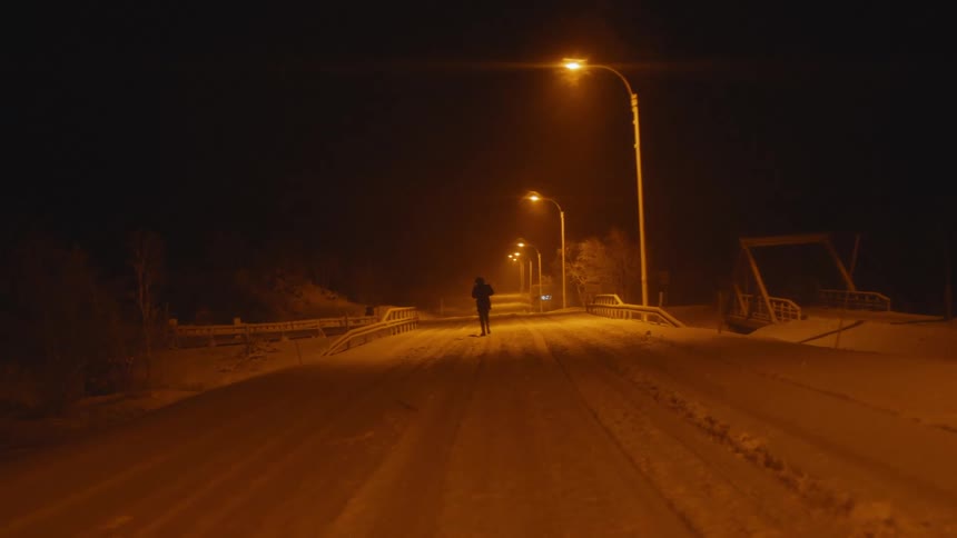 Pedestrian walks down a snowy road under the streetlights at night