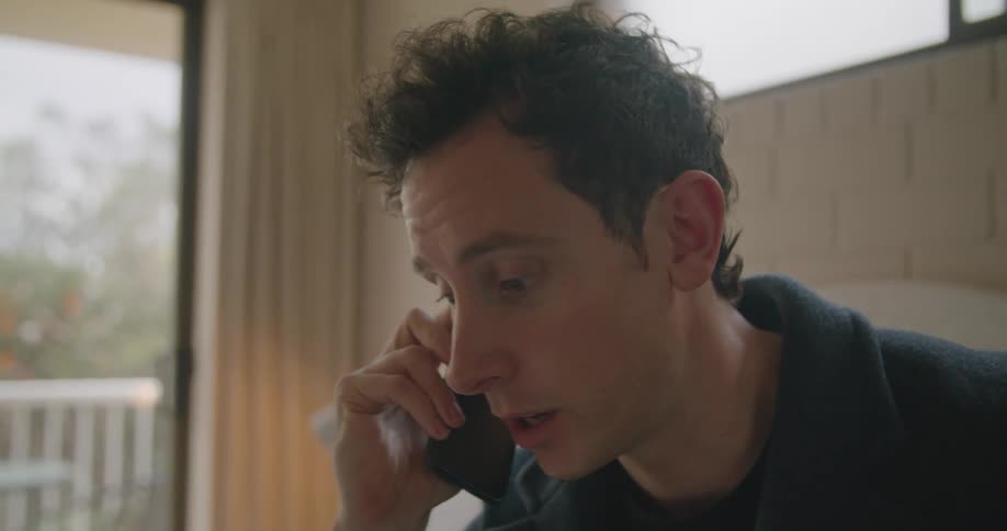 A man talking on the phone while using a laptop on a bed