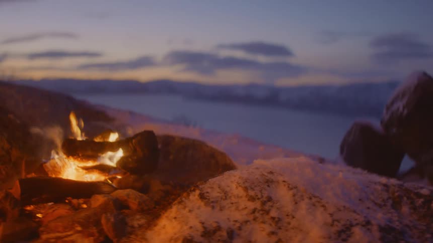 Camp fire burns at dusk with clouds in the sky and snow on the ground