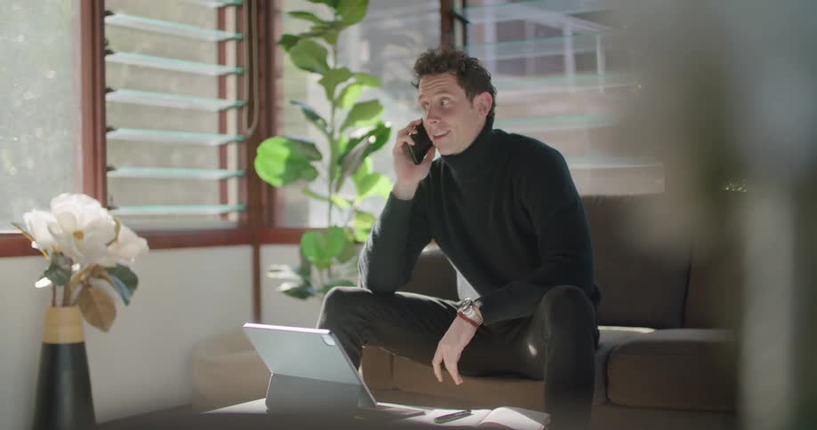 Man sits in living room talking on mobile phone and viewing tablet