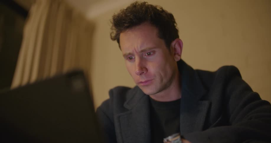 A man in a room using a laptop with a wall and window in the background