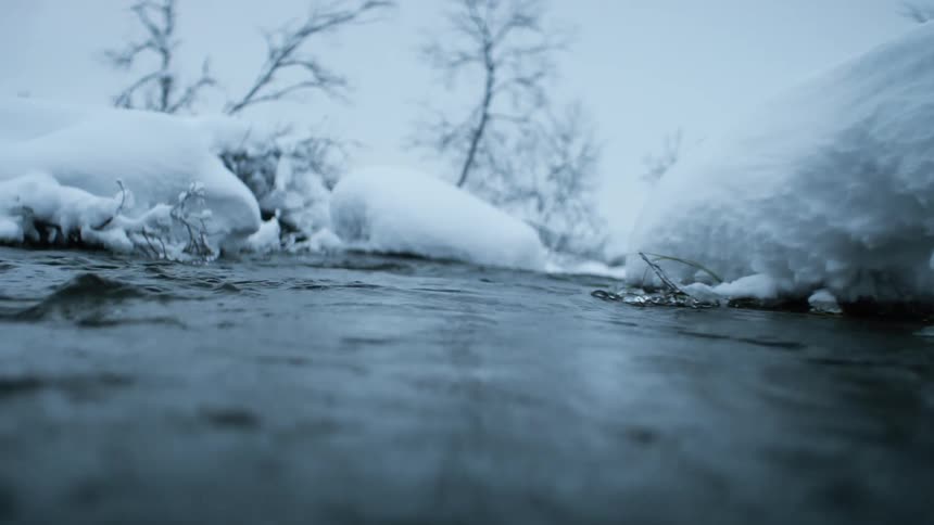 A river flows steadily past a snow covered landscape as leafless trees stand nearby