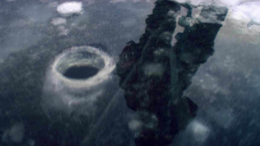 A fisherman stands next to a fishing hole drilled into a ice covered lake