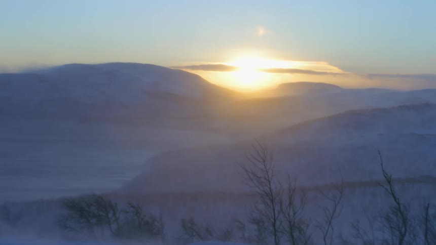 Sun rising in the horizon over a snowy mountain range while wind blows snow and tree branches