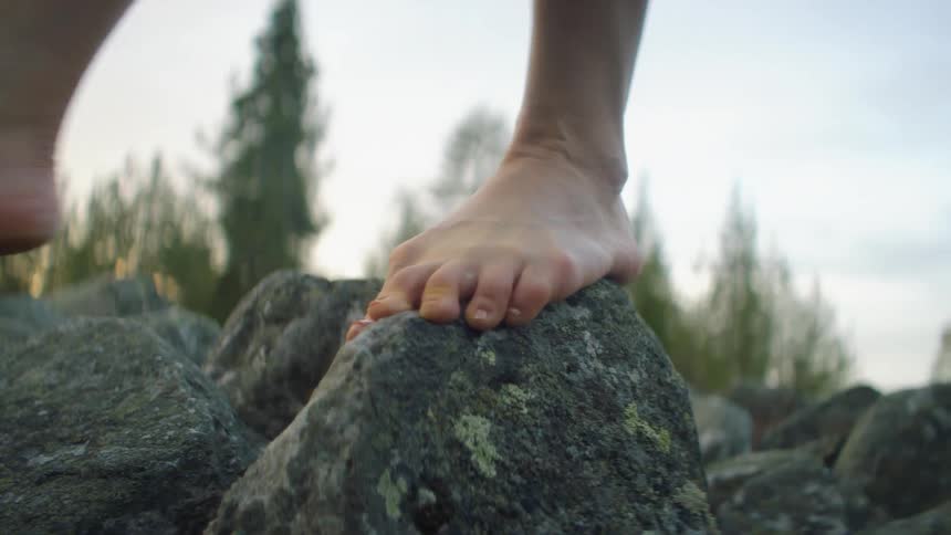 A person barefooted walking on rocks with trees the sky and clouds in the background