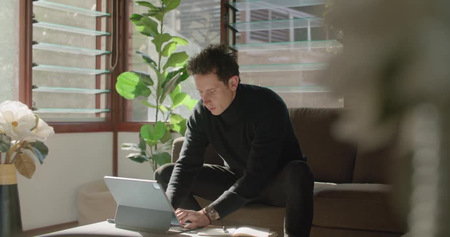 A man writes in a notebook then searches on his tablet while sitting at home on a couch