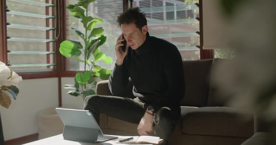 Man sitting in a couch ends a call on the smartphone and keeps working on a laptop
