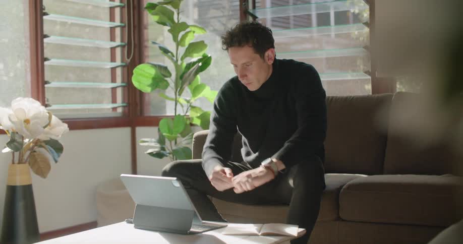 A man sitting on a couch in a bright office makes notes then types on a laptop