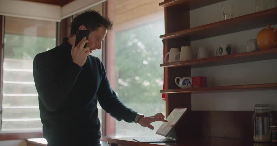 A man in a turtleneck sweater talking on a phone while looking at a laptop at home