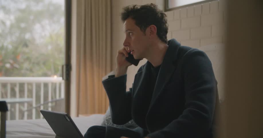 Man with blazer jacket sitting on bed talks on smartphone and uses tablet