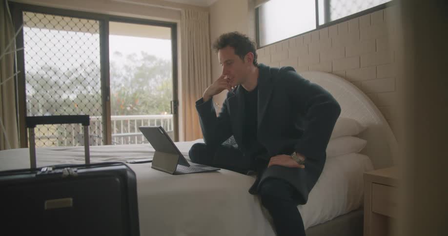 A man is talking on a laptop and sitting on a bed indoors
