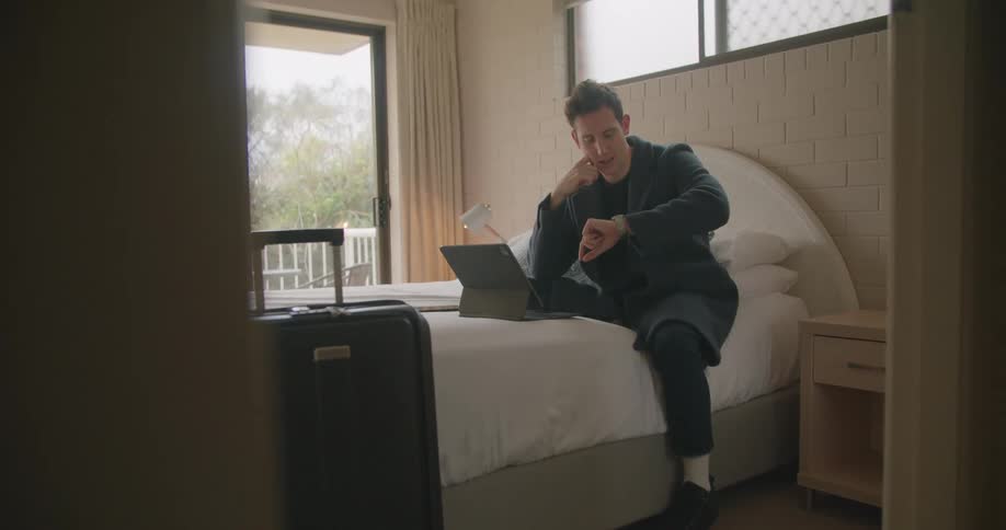 Man sitting over a bed in a hotel room talking on a video conference on a laptop