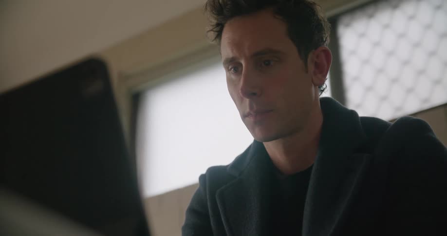 A man in a room using a laptop with a wall and window in the background