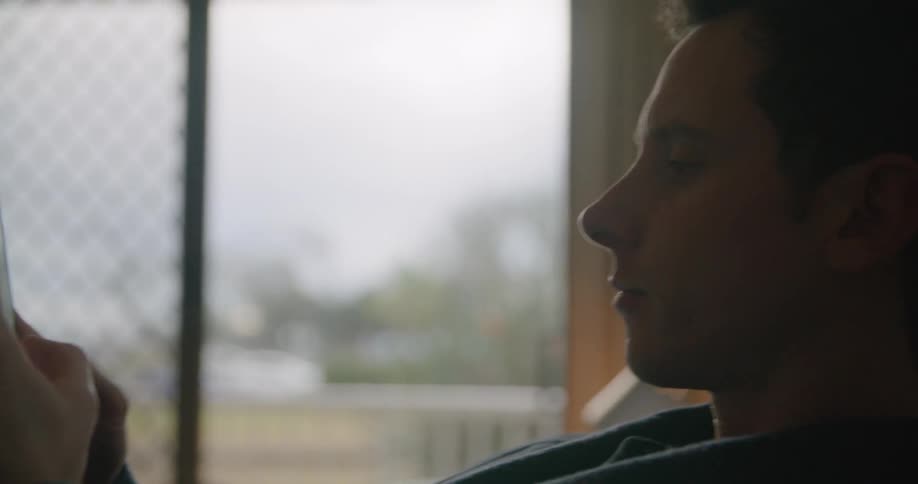 A man sitting up against a bed headboard texting on a smartphone during the day