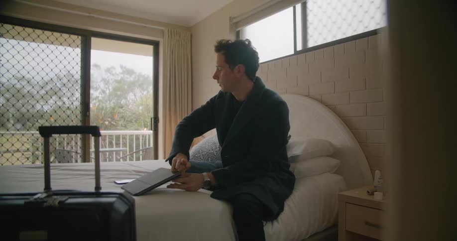A man in a room sitting on a bed looking at a smart phone than opening up a tablet