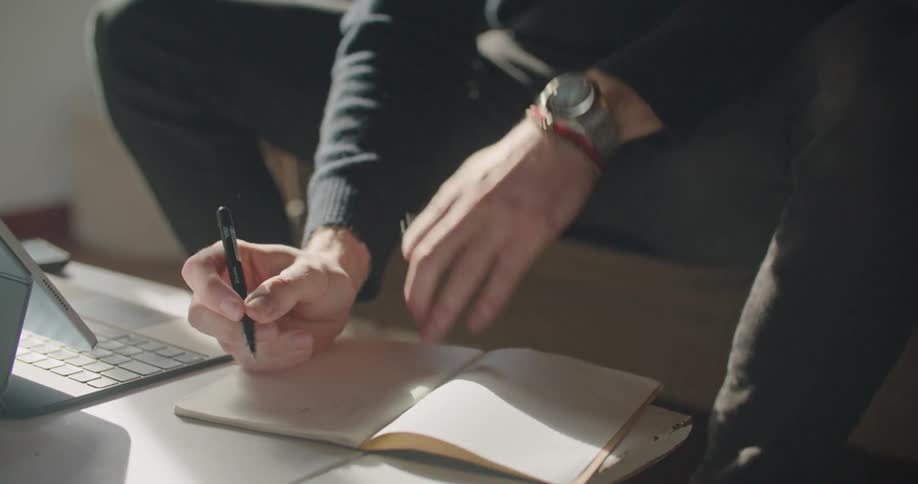 Man in a black turtleneck sweater and pants sits on couch and takes notes by looking at tablet