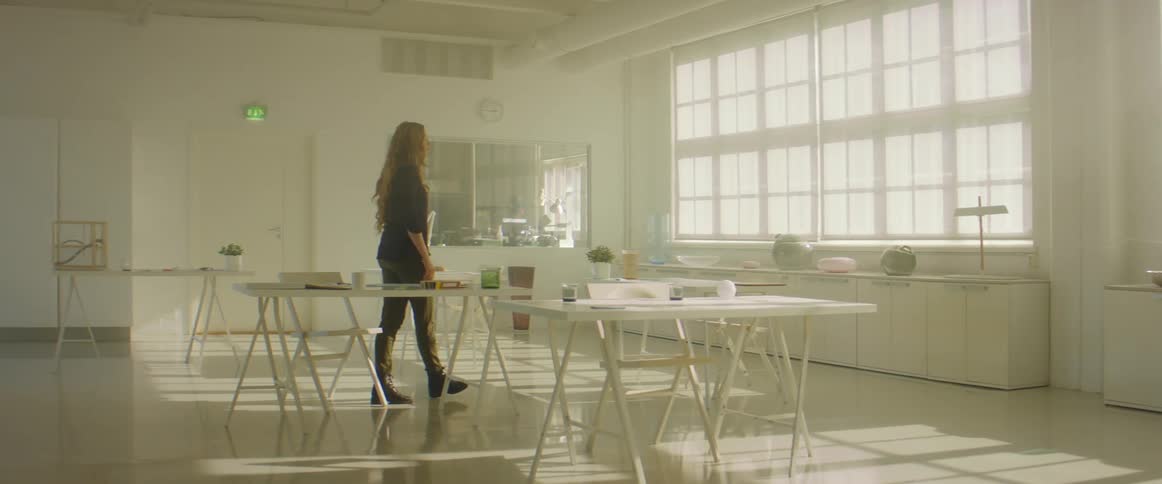 A woman in a room sitting on a chair at a desk then standing up with a wall and windows in the background