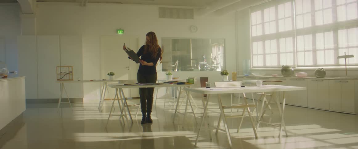 A woman holding a book of designs looking over work on a table inside of a room at work during the day