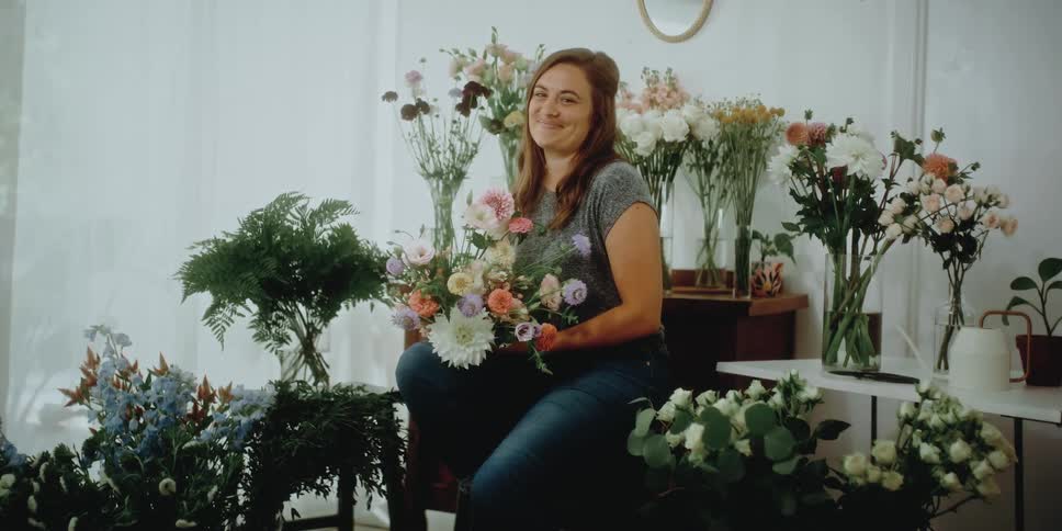 A female florist smiles as she sits and poses while surrounded by many floral arrangements