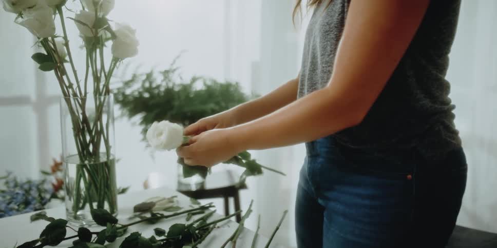 A woman plucking the leaves off of white roses on a table to be placed in a vase with water on a table indoors during the day