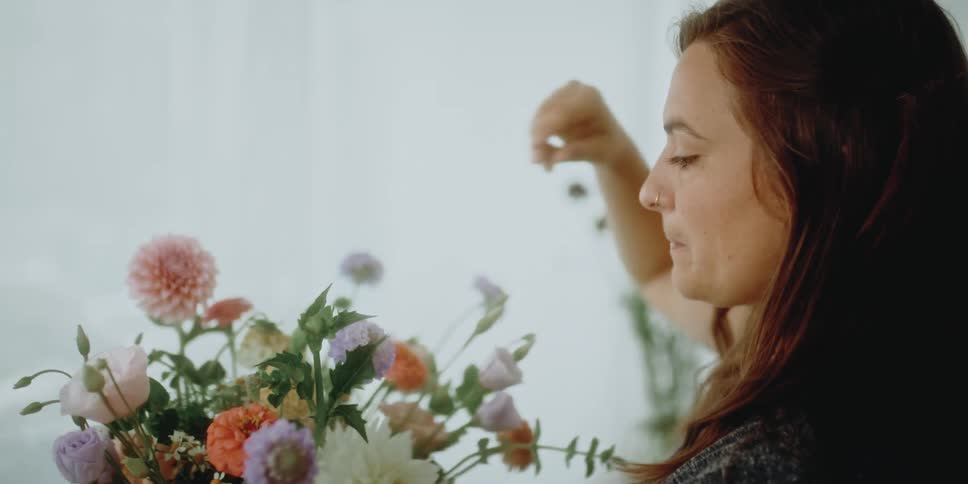 A woman in a room arranging flowers with a wall and plants in the background