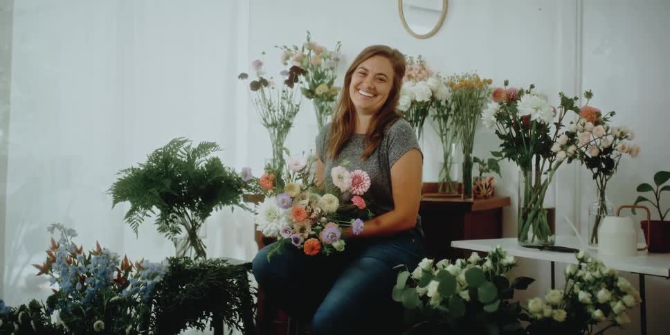 Happy florist surrounded by flowers sits with a flower bouquet in her hands