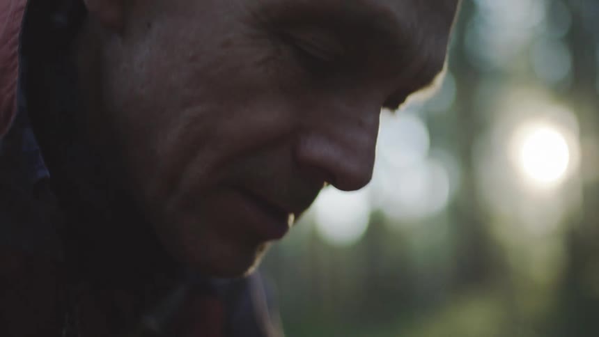 An elderly man bends over to pick something up in a forest