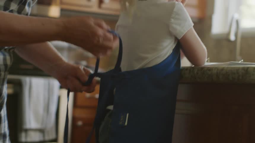 A little girl stands in a kitchen while a family member ties a blue apron around her back during the day