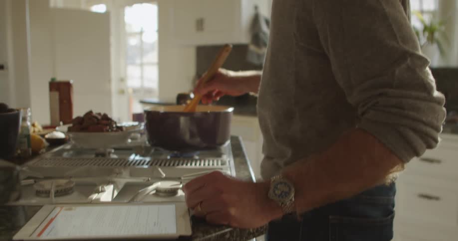 A man reads a recipe on his tablet while stirring food on the stove