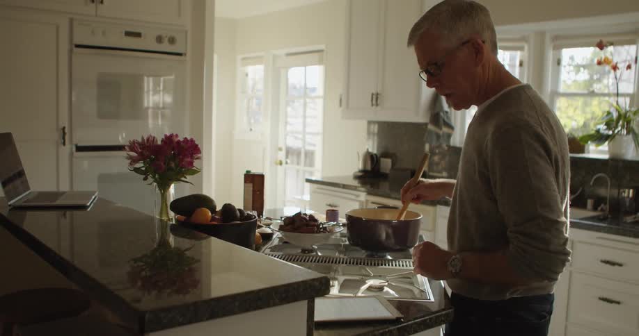 Man stirs food in a pot with a wooden spoon while looking at a recipe on the counter