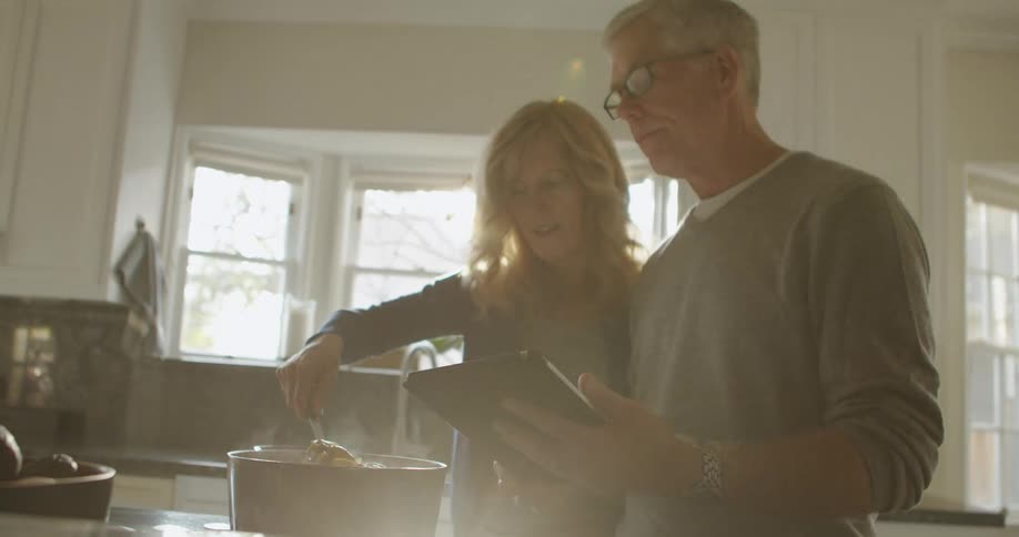 Man with glasses and grey sweatshirt holds tablet while blonde woman cooks in the kitchen