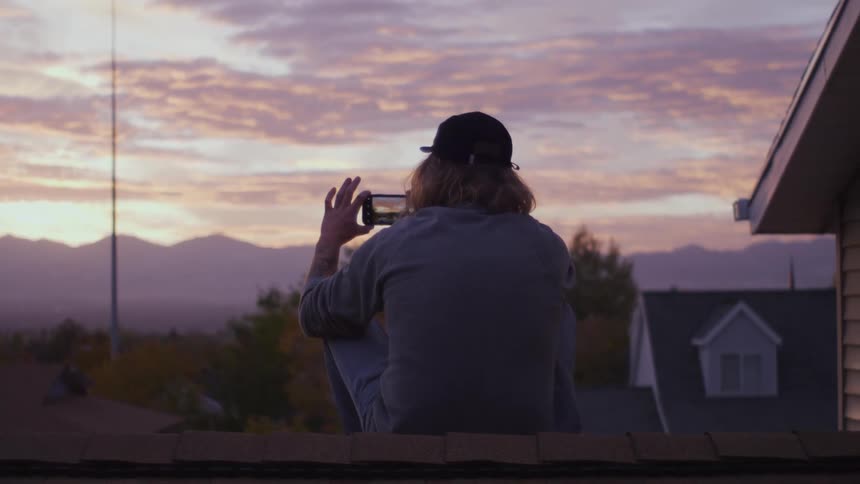 Man filming the sunset with a smartphone while sitting on rooftop