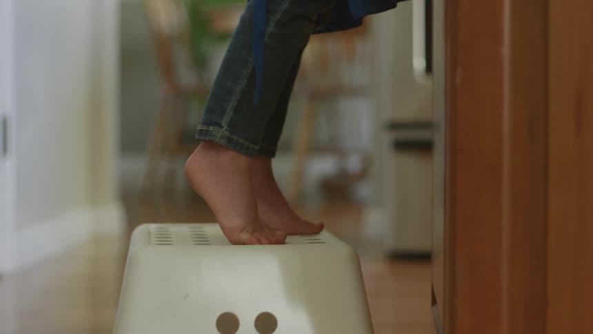A woman standing on a stool is standing on her toes reaching for something in a residential kitchen