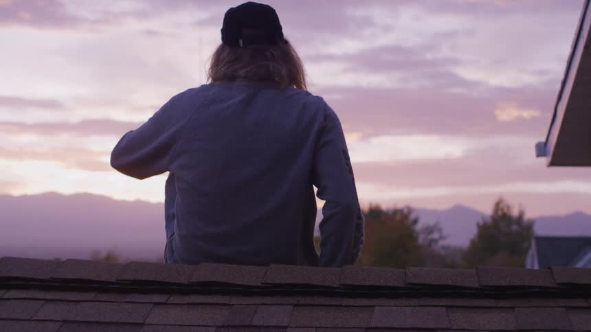 Man wearing a hat sits on the peak of a roof and looks at the mountains in the distance