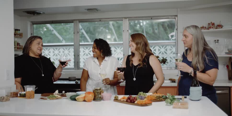 A group of women is standing by a counter covered with food and wine
