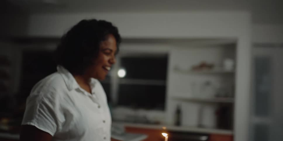A woman at home carries a cake with a candle to three other women sitting at the table