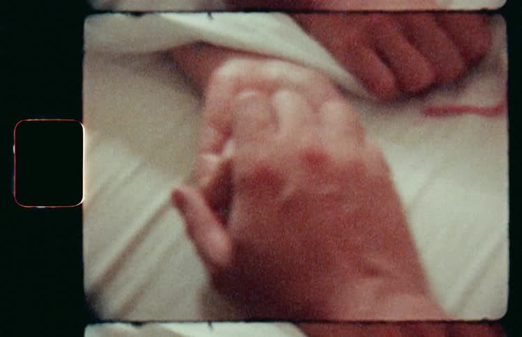 As one person lays in a bed under the sheets another person holds and caresses their hand