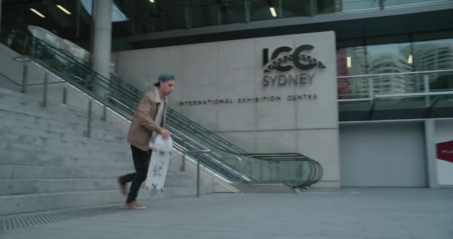 A man walks down a set of stairs before getting on a skateboard and skating down a walkway at the bottom of the staircase