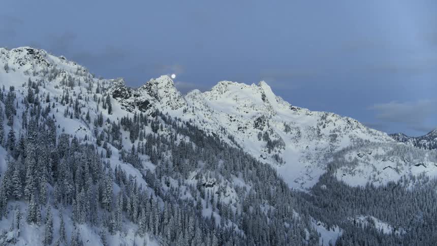 A bright white moon emerges from behind a jagged and snow covered mountain ridge
