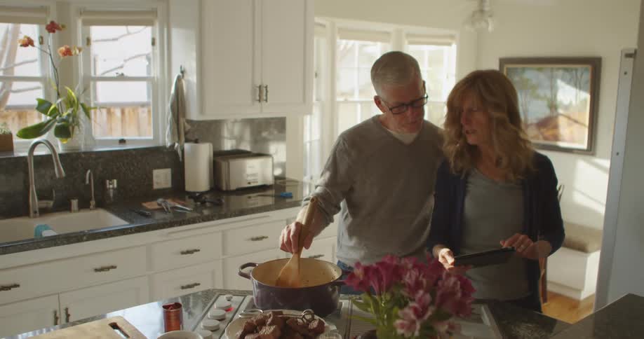 Older couple stand in kitchen cooking while consulting tablet