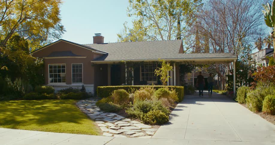 A house is shown with a lot of landscaping while two men walk into the carport
