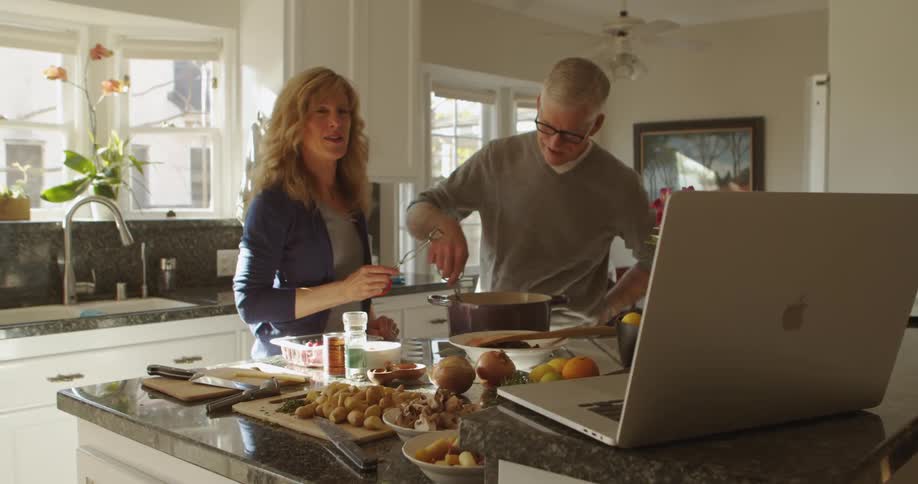A man and a woman are in a kitchen watching something on a laptop as they cook a meal