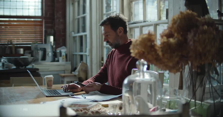 Man sitting at a desk working on a laptop while using a smartphone